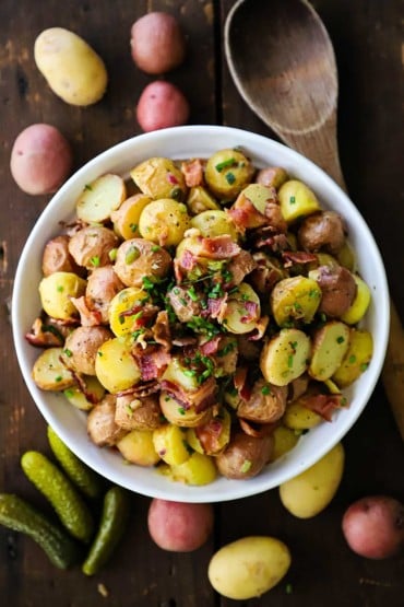 An overhead view of a white bowl holding a large serving of German potato salad with a few whole baby potatoes and petite dill pickles surrounding the bowl.