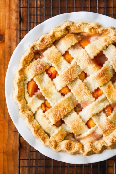 An overhead view of a peach pie with a pastry lattice topping all sitting on a baking wire on a wooden cutting board.