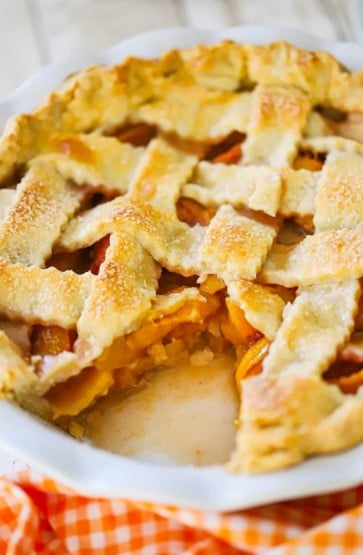 A straight-on view of a homemade peach pie in white pie dish and there is a slice missing, revealing the peach filling.