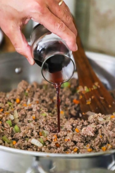 A person pouring red wine from a small carafe into a large silver skillet filled with cooked ground beef, Italian sausage, and sautéed vegetables.