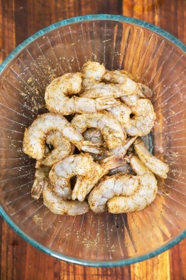 A glass bowl of large uncooked shrimp that has been tossed with Cajun seasoning and olive oil.