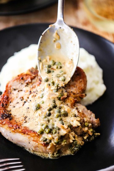 A person using a silver spoon to drizzle on a creamy caper sauce over a seared pork chop on a black plate with mashed potatoes.