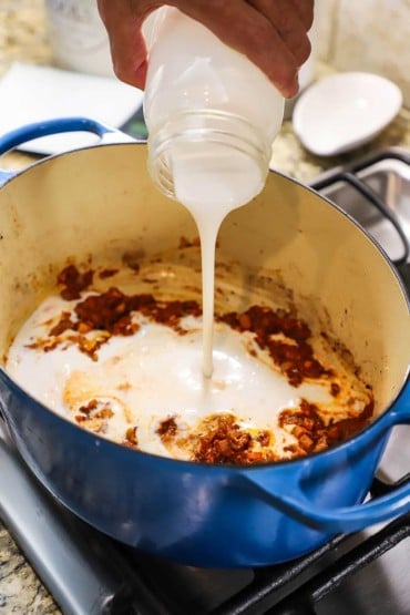 A person pouring unsweetened coconut milk from a jar into an oval Dutch oven filled with sautéd onions coated with red curry paste.