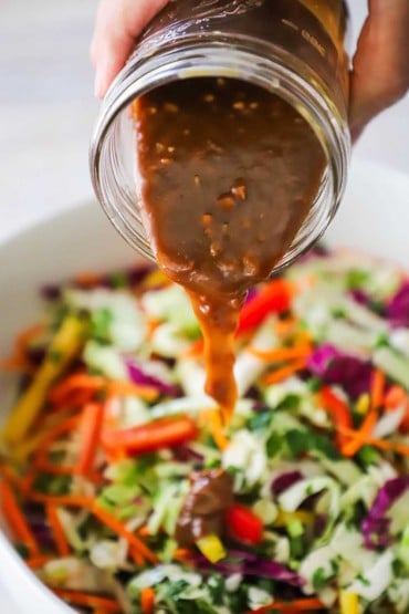 A person pouring a peanut dressing from a small glass jar into a white bowl filled with an Asian slaw.