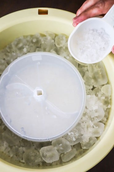 A person using a plastic half-cup measuring cup to transfer rock salt into the basin of an electric ice cream maker that is full of ice surrounding a metal canister filled with churning cream.