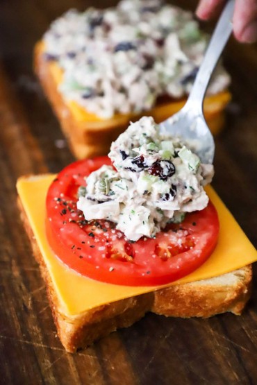 A person using a spoon to place homemade tuna salad on top of a slice of tomato that is sitting on a piece of bread with a slice of cheese on it.
