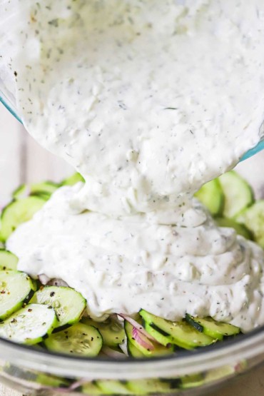 A large amount of homemade tzatziki sauce being poured from a medium-sized glass bowl into another bowl filled with sliced cucumbers and thinly sliced red onion.