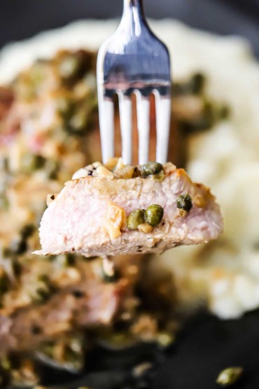 A piece of pork chop being held up by a fork with capers on it.
