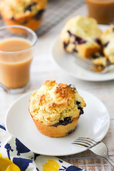 A lemon and blueberry jumbo muffin that is sitting on a small white plate next to a glass mug full of coffee and cream.