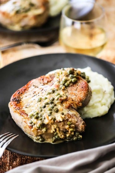 A front-on view of a black plate filled with mashed potatoes and a seared pork chop with caper sauce on top of it.