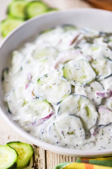 A close-up side view of a white serving bowl filled with tzatziki cucumber salad with fresh cucumber slices sitting nearby.