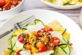 A straight-on view of two white bowls filled with a serving of zucchini and yellow squash salad topped with a cherry tomato relish, pine nuts, and shave parmesan cheese.