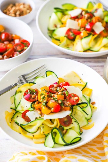 A straight-on view of two white bowls filled with a serving of zucchini and yellow squash salad topped with a cherry tomato relish, pine nuts, and shave parmesan cheese.