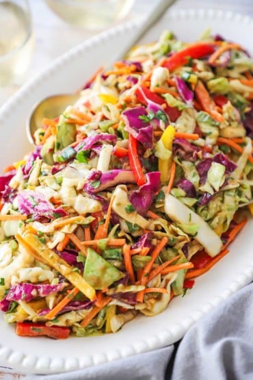 A close-up view of an Asian slaw with Peanut Ginger Dressing on a white platter with a golden serving spoon inserted into the side.