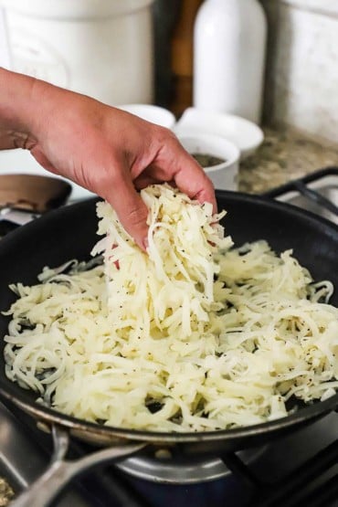 A person dropping a handful of shredded partially cooked russets potatoes in a large non-stick skillet on a stove.