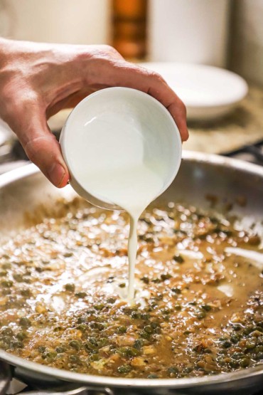 A person pouring heavy cream from a small white bowl into a large stainless steel skillet filled with a caper sauce.