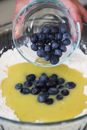 A person transferring fresh blueberries from a small glass bowl into a large glass bowl filled with a flour mixture and egg and lemon batter in the middle of it all.