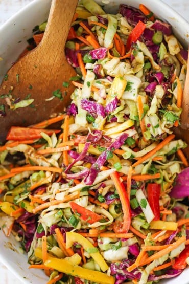 An overhead view of a slaw made with diced red and green cabbage, red and yellow bell peppers, and scallions in a bowl with two wooden spatulas off to the side.