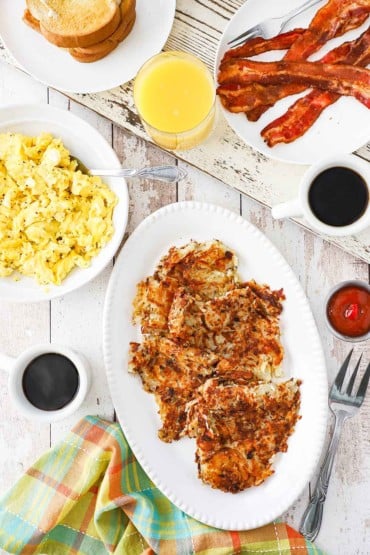 An overhead view of a platter of crispy hash brown next to a bowl of scrambled eggs, two cups of black coffee, a glass of orange juice, and a plate of cooked bacon.