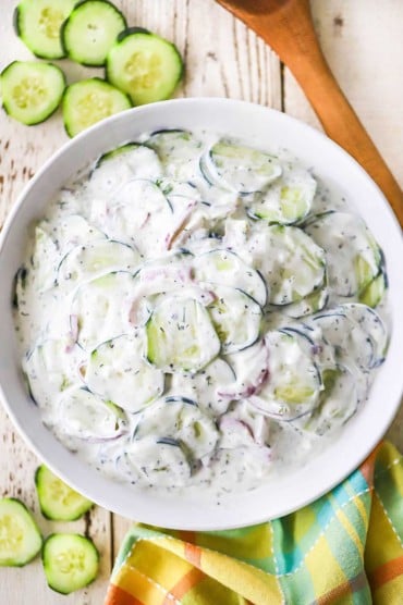 An overhead view of a white bowl filled with Tzatziki Cucumber Salad that is flanked by a wooden spoon and freshly sliced cucumbers.