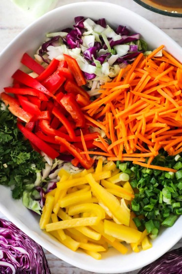 An overhead view of a large square serving bowl filled with piles of shredded carrots, cabbage, red and yellow bell peppers, herbs, and chopped scallions.