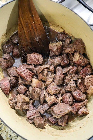 An overhead view of bite-sized pieces of chuck roast that are being seared in an oval Dutch oven with a wooden spatula off to the side.