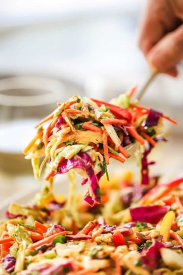 A person lifting a large serving spoon of Asian slaw with peanut ginger dressing from a bowl of the same.