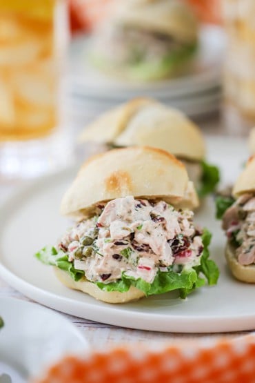 A straight-on view of a white circular platter filled with dinner rolls that have been cut in half and filled tuna salad on a layer of green leaf lettuce.