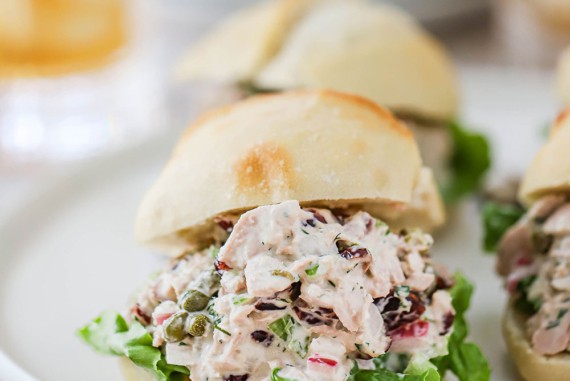 A straight-on view of a white circular platter filled with dinner rolls that have been cut in half and filled tuna salad on a layer of green leaf lettuce.