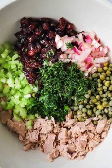 An overhead view of a bowl filled with small piles of chopped tuna, celery, dried cranberries, chopped onion, capers, and fresh herbs.
