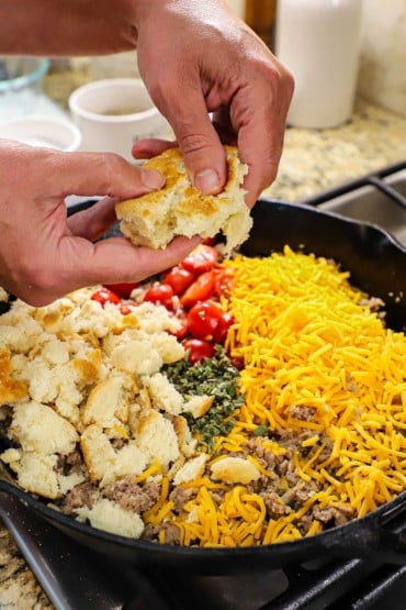 A person using two hands to crumble buttermilk biscuits into a large cast-iron skillet filled with cooked sausage, shredded cheese, chopped sage, and cherry tomatoes.