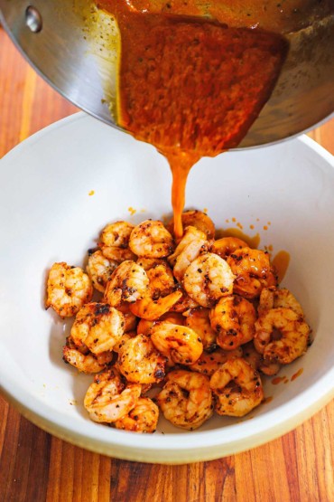 Buffalo sauce being poured from a steel saucepan into a ceramic bowl of grilled shrimp.