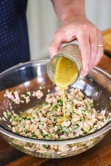 A person pouring a mustard vinaigrette from a small glass jar into a large glass bowl filled with an albacore tuna and white bean salad.