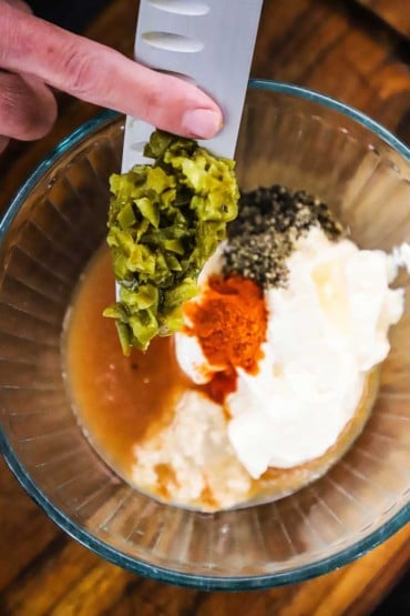 A person scraping chopped pickled jalapeno peppers into a glass bowl filled with mayonnaise, cayenne pepper, black pepper, and apple cider vinegar.