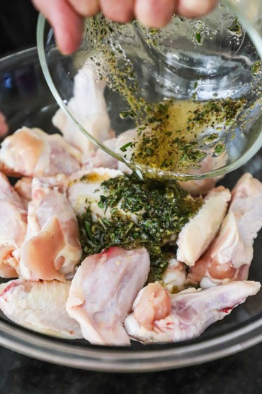 A person pouring a marinade of oil, garlic, and herbs over a pile of uncooked chicken wings in a glass bowl.