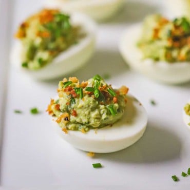 A close-up view of an avocado deviled egg sitting on a white platter with snipped chives and bread crumbs on top.