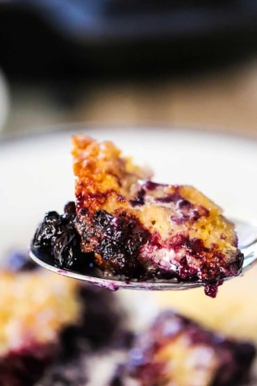 A spoon holding up a helping of blueberry cobbler over a bowl of the same.
