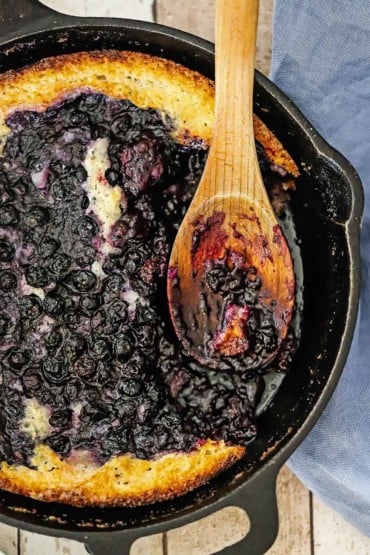 An overhead view of a cast-iron skillet containing blueberry cobbler with a portion of the cobbler missing and a wooden spoon inserted into the side.