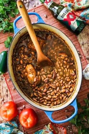 An overhead view of a large oval Dutch oven filled with borracho beans with a wooden spoon inserted into the middle of them.