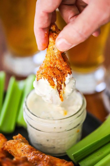 A person pulling a Buffalo wing leg from a small jar filled with blue cheese dressing and there is dressing from the wing.