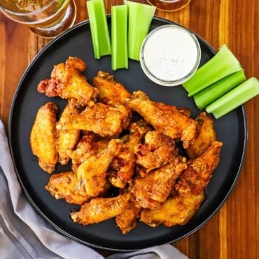 An overhead view of pile of classic Buffalo wings on a black circular plate also holding celery sticks and a small jar of blue cheese dressing.