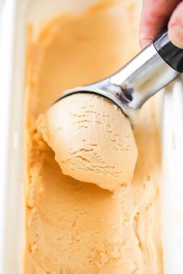 A person using an ice cream scooper to roll a scoop of salted caramel ice cream in a rectangular ice cream container.