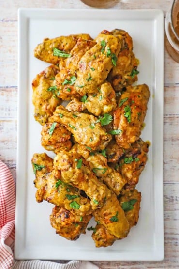 An overhead view of a pile of grilled curry chicken wings topped with chopped cilantro on a white rectangular platter.