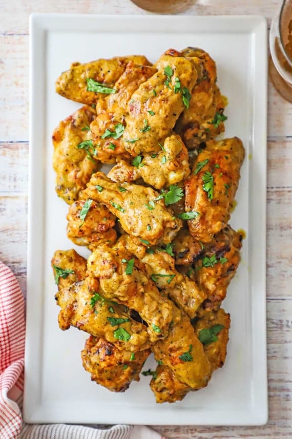 An overhead view of a pile of grilled curry chicken wings topped with chopped cilantro on a white rectangular platter.