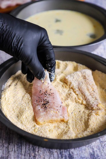 A person a wearing black nitrate glove dredging a fillet of catfish through a pan filled with a cornmeal and flour mixture.