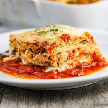 A white dinner plate with a square serving of eggplant parmesan sitting in thin layer of marinara sauce.