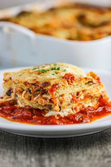 A white dinner plate with a square serving of eggplant parmesan sitting in thin layer of marinara sauce.