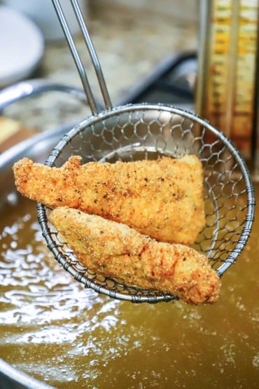 Two fried catfish fillets resting in a metal spider over a skillet filled with hot oil.