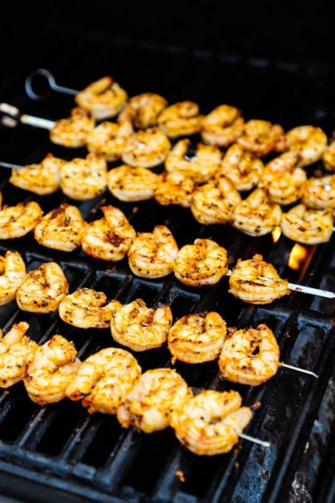 Six rows of shrimp on metal skewers on the grates of a gas grill with a flame visible below.