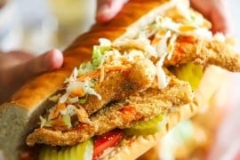 A person holding a large fried catfish po-boy that is stuffed with crispy fillet of catfish, coleslaw, hot sauce, pickles, and tomatoes.